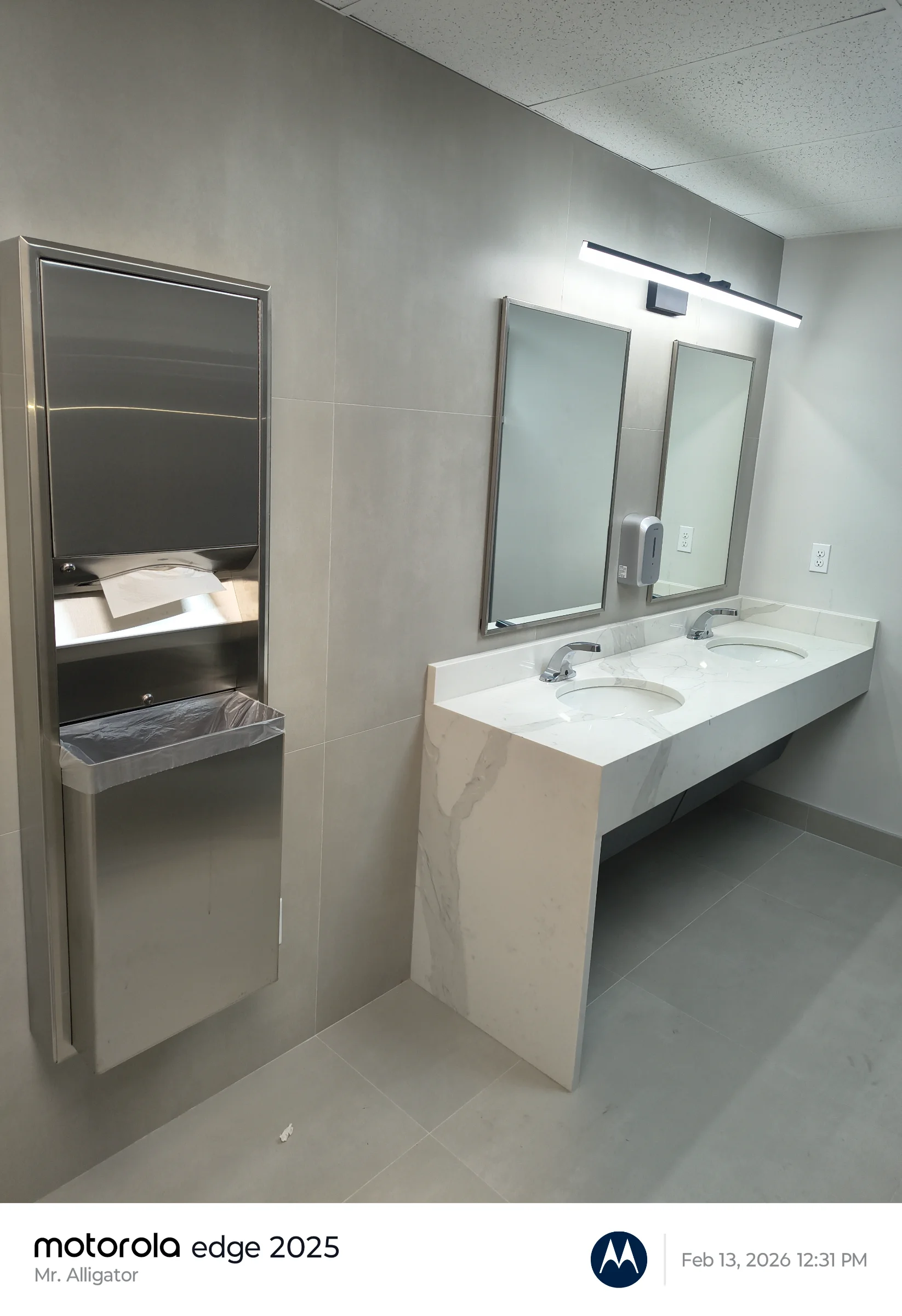 Completed double-sink quartz countertop vanity with undermount basins, touchless faucets, wall mirrors, and recessed stainless steel paper towel dispenser.