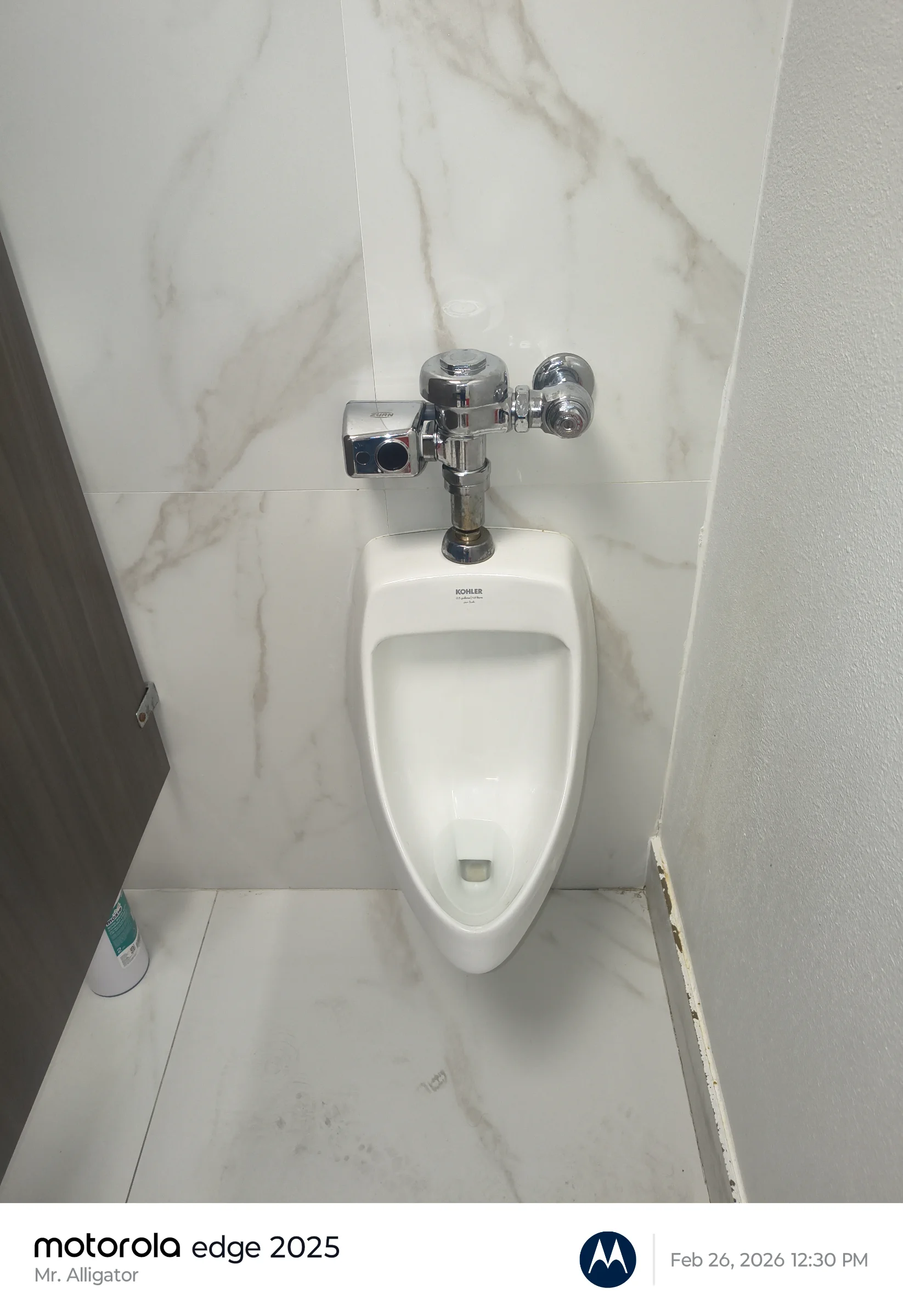 Touchless wall-mounted Kohler commercial urinal installed against marble-patterned porcelain wall tiles.